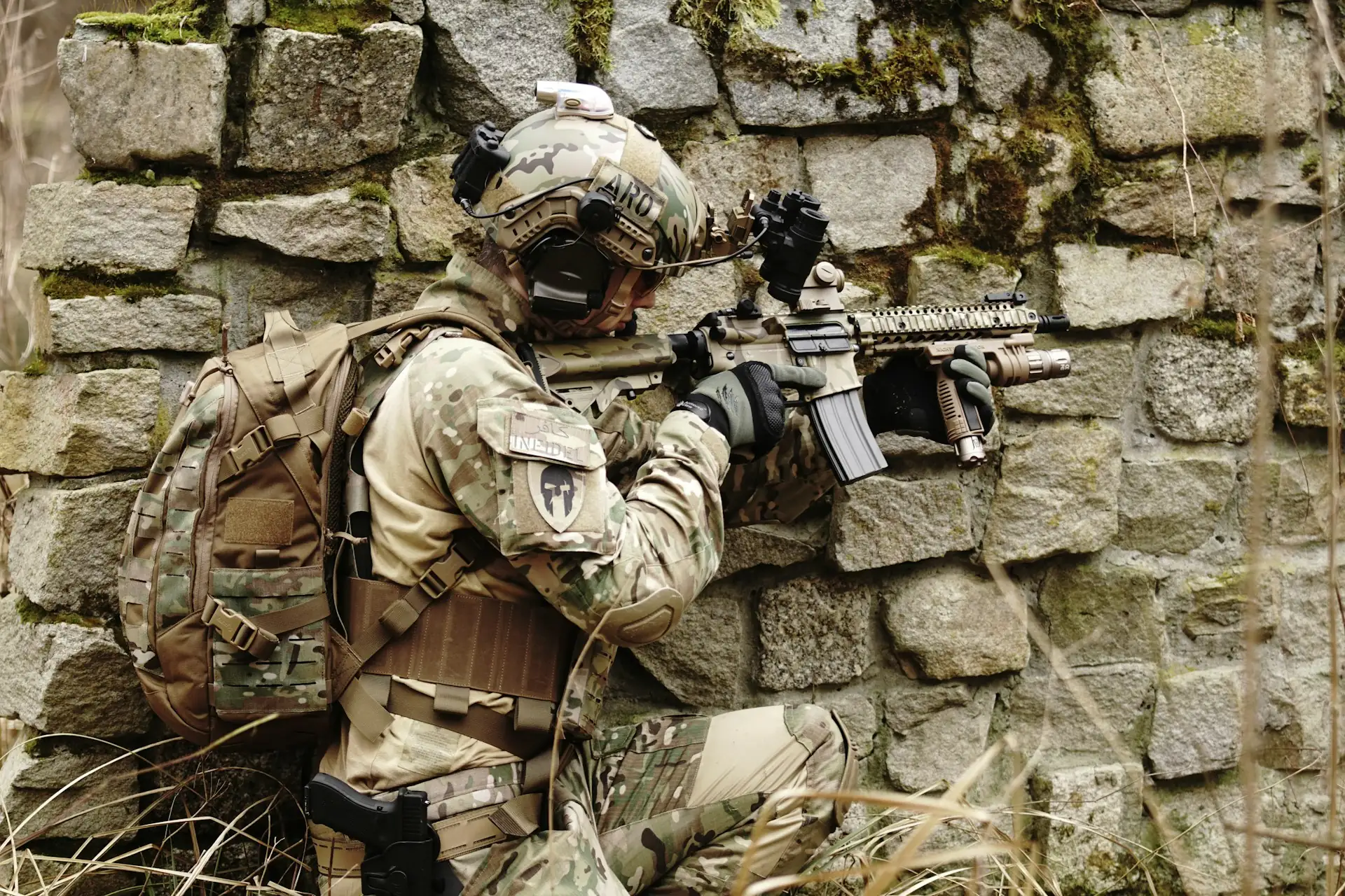 Soldier in field gear aiming a rifle from cover behind a stone wall
