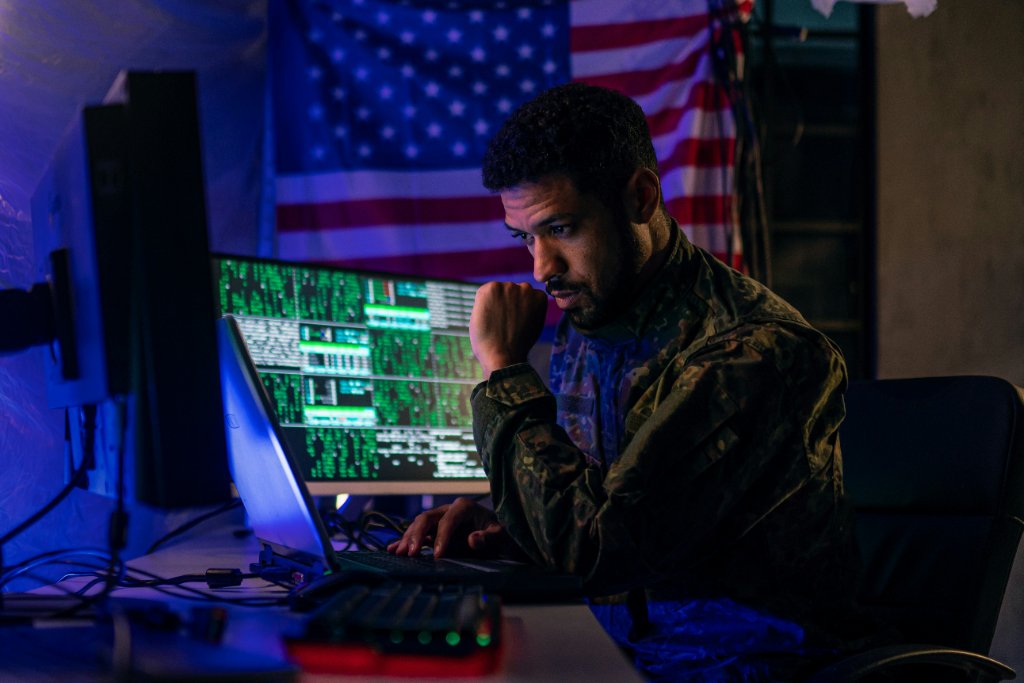 Analyst in a camouflage uniform reading network data on two screens, a U.S. flag on the wall behind
