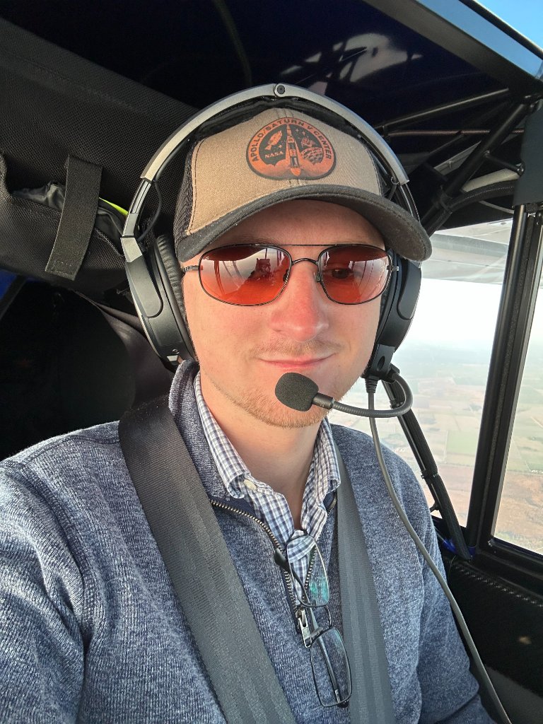 Cody Franklin in the cockpit of a light aircraft, headset on, farmland below